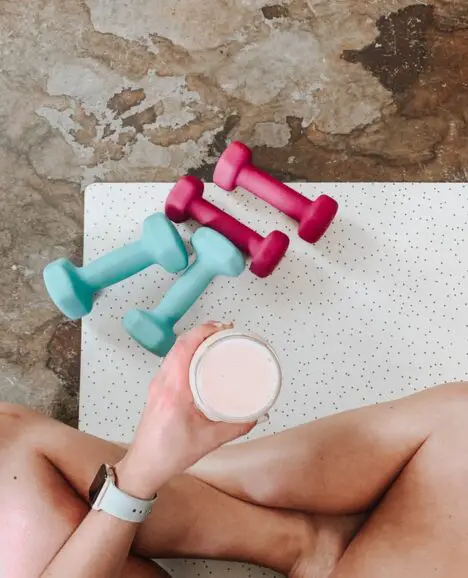 A woman holding a protein shake next to weights